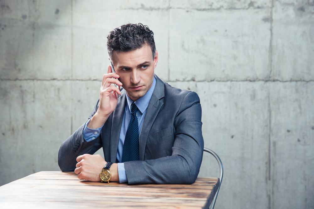 Pensive businessman sitting at the table and talking on the phone over concrete wall. Looking away